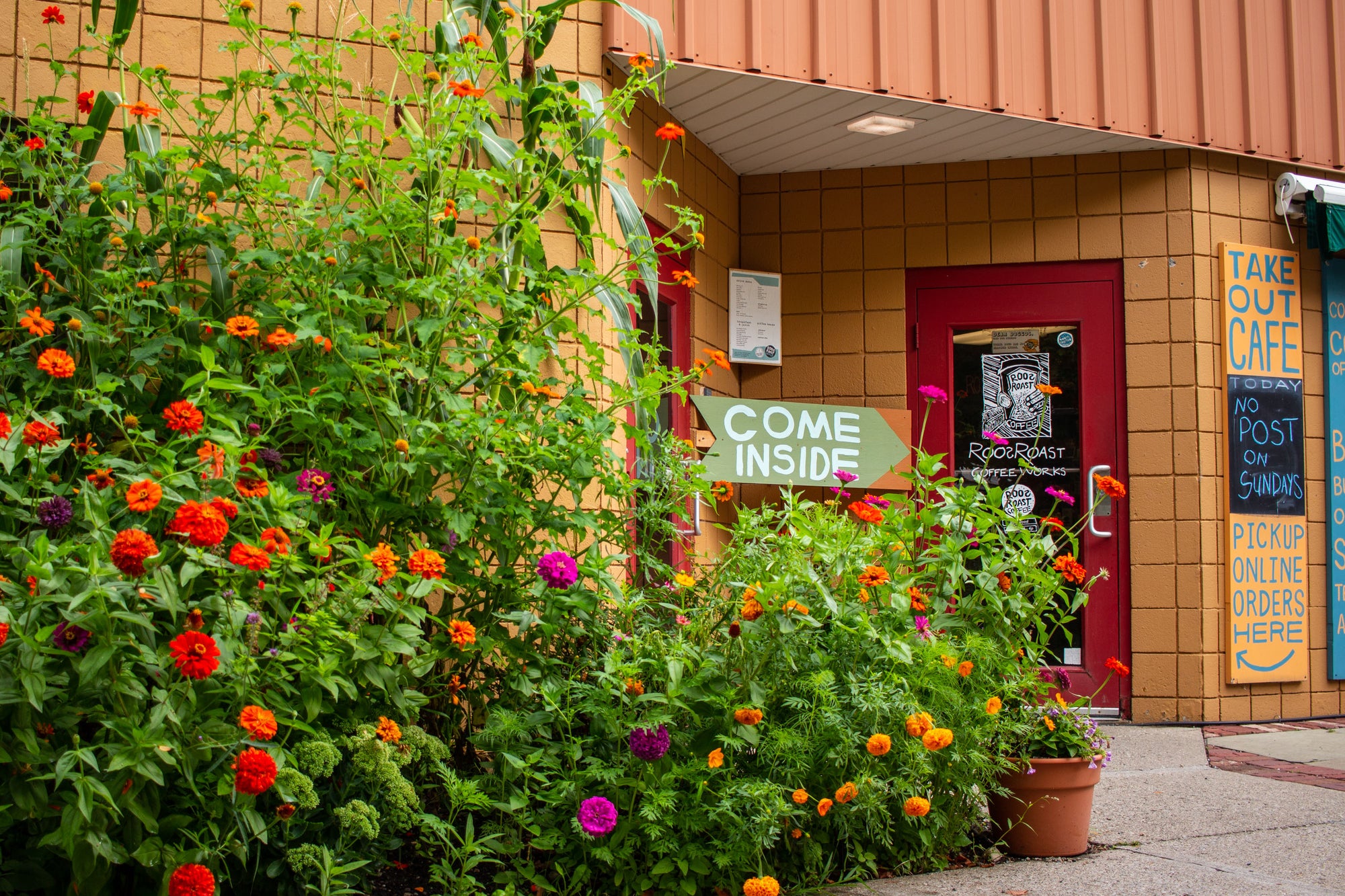 A storefront entrance with a red door, tan brick wall, and abundant green foliage in the foreground featuring orange, red, and pink flowers. A sign reads “COME INSIDE,” and a vertical blue sign on the right advertises a cafe and post office.
