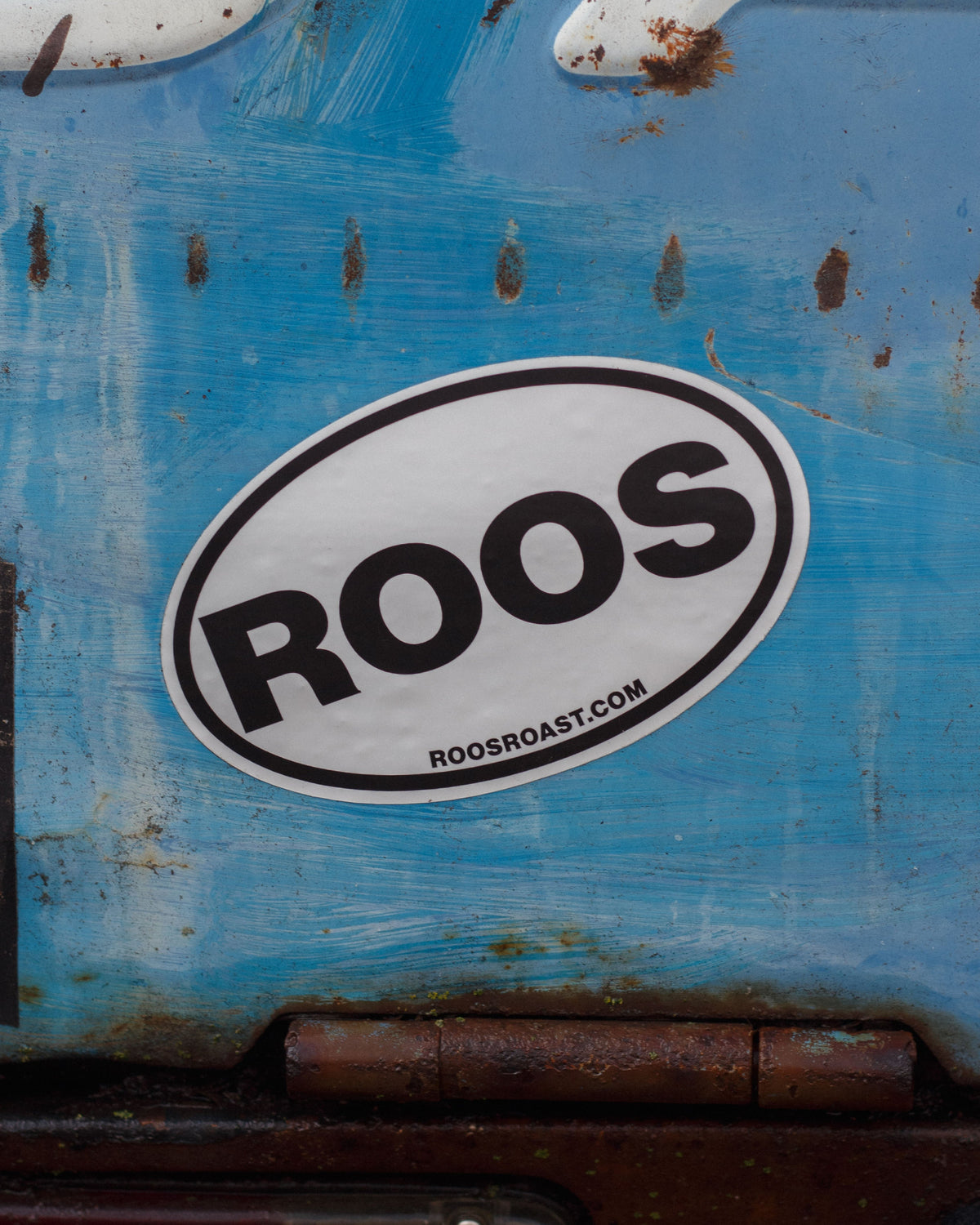 Sticker with 'ROOS' on a blue surface with rust and wear.