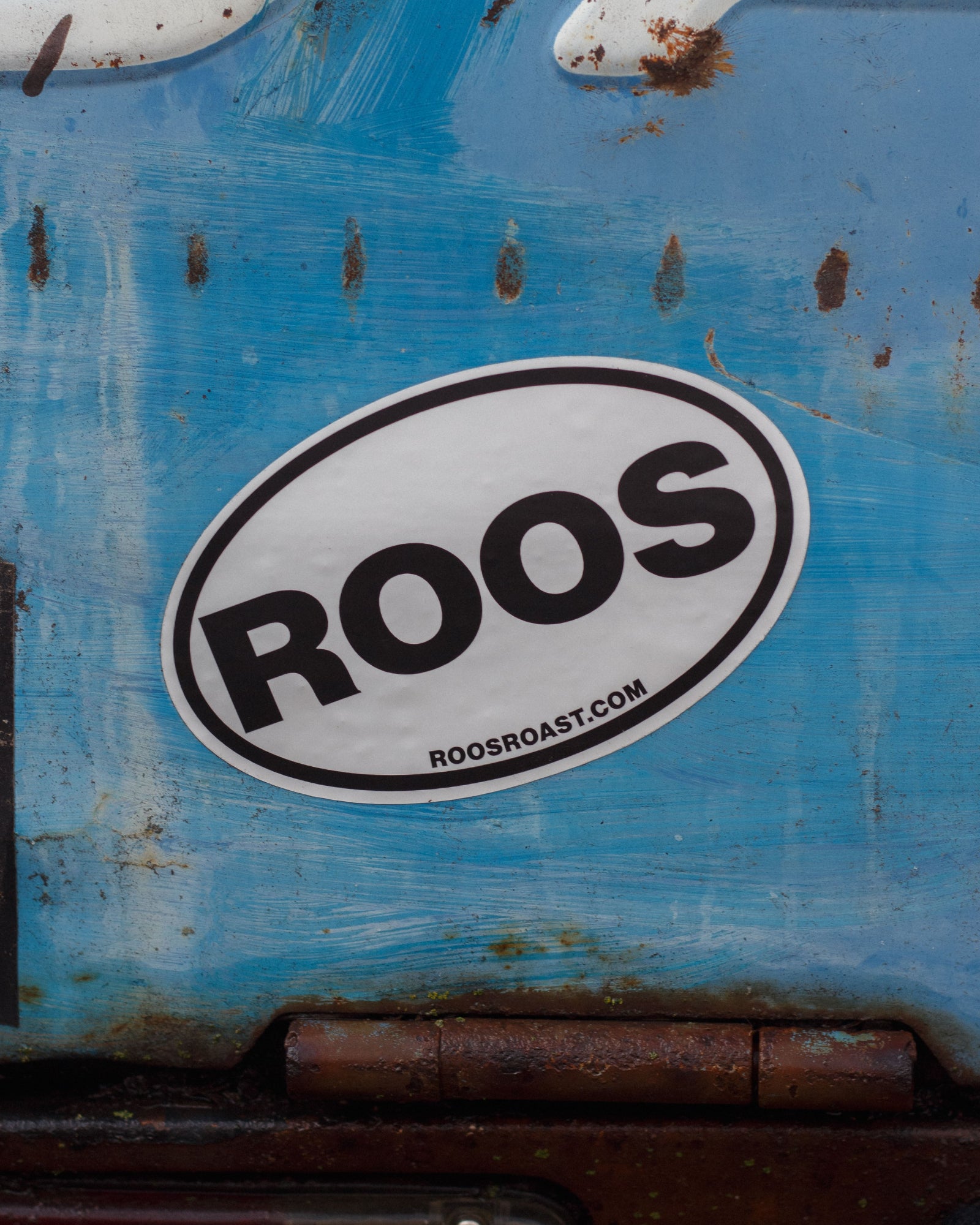 Sticker with 'ROOS' on a blue surface with rust and wear.
