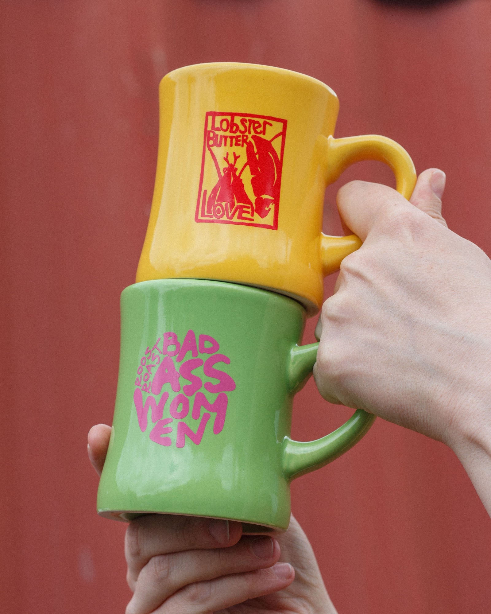 Two ceramic mugs stacked on top of each other, held by hands against a red background. The top mug is yellow with a red 'Lobster Butter Love' graphic, and the bottom mug is green with pink 'BADass WOMEN' text.