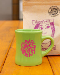 Green mug with 'Bad Ass Women' text in pink on a wooden surface with a coffee bag in the background.