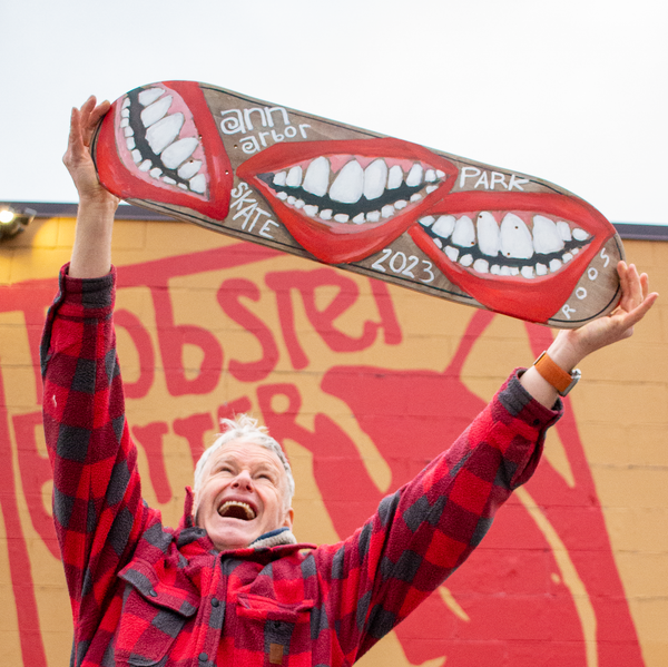 John Roos holds up a hand painted skate board in front of the lobster butter love mural 