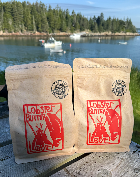 Two packages of 'Lobster Butter Love' on a wooden surface with a scenic background of water and trees.