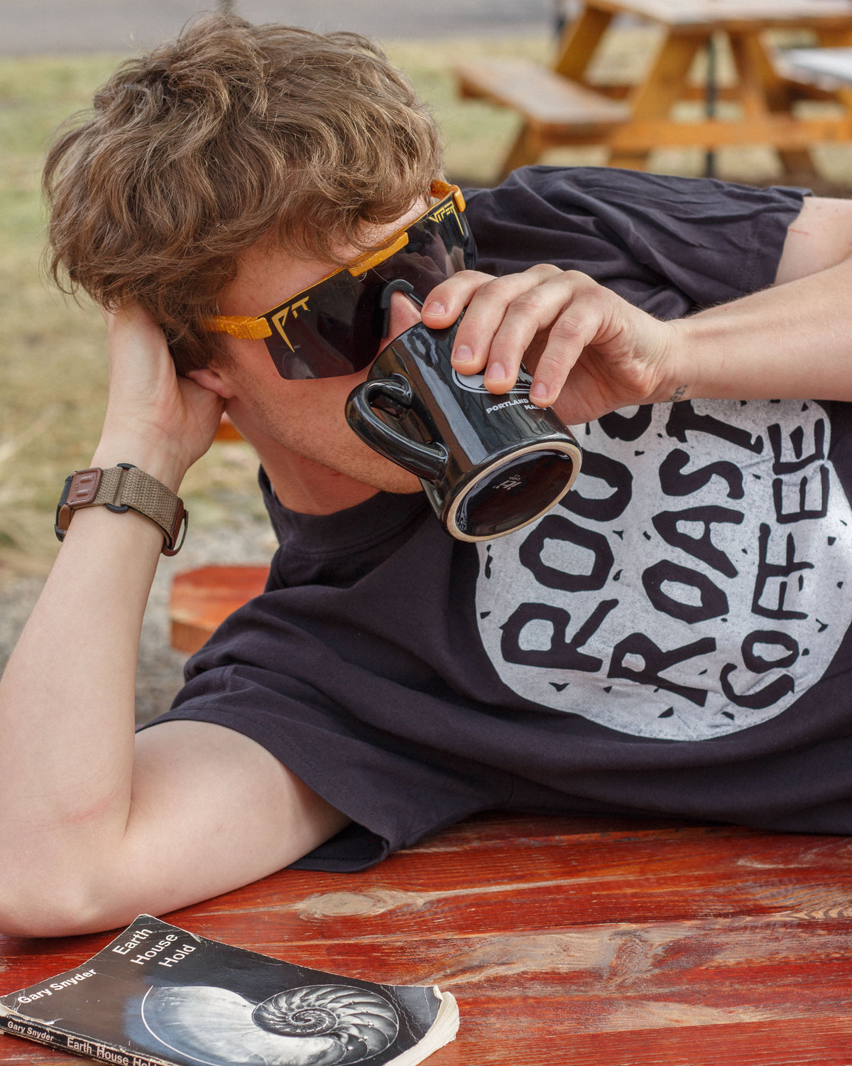 Person wearing sunglasses and a 'Rooftop Coffee' t-shirt, drinking from a mug outdoors.