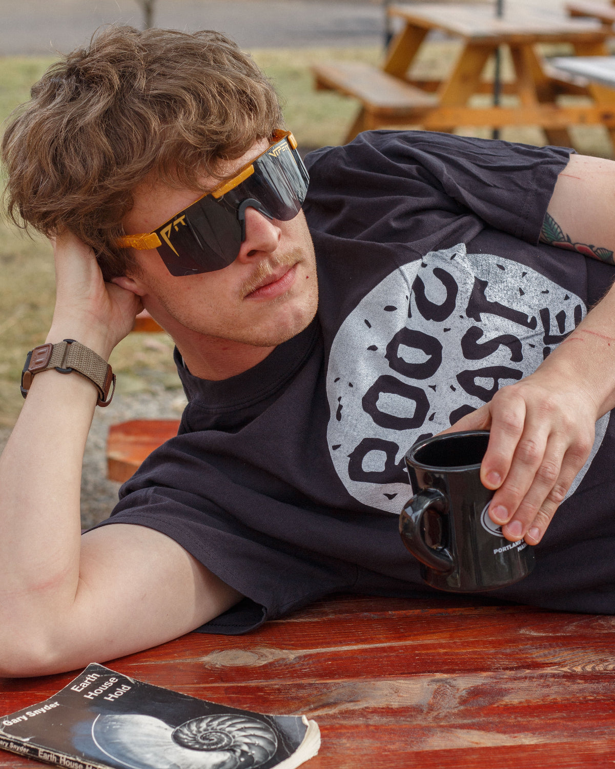 Person wearing sunglasses and a black t-shirt with a graphic design, sitting at an outdoor table.