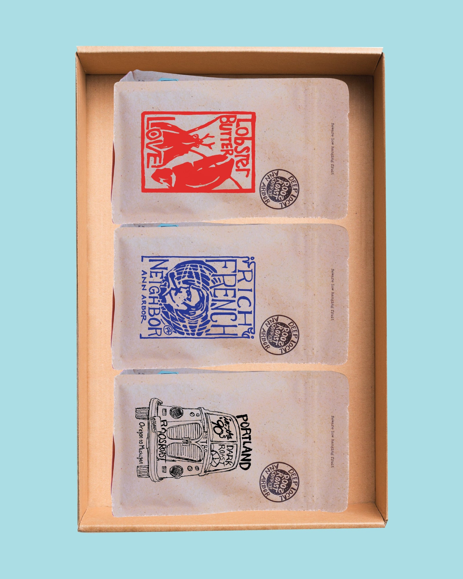 Three small paper mailers or envelopes stacked inside a light wood box, each printed with a different colorful stamp-style graphic and text including 'LOBBER LOVE', 'FRENCH NEIGHBOR', and 'PERSHING'.