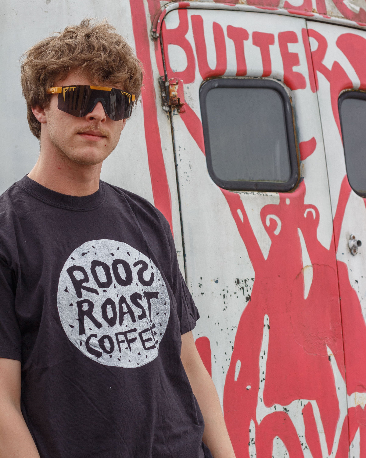 Person wearing a black t-shirt with 'Roos Roast Coffee' logo in front of a vehicle with red graphics.