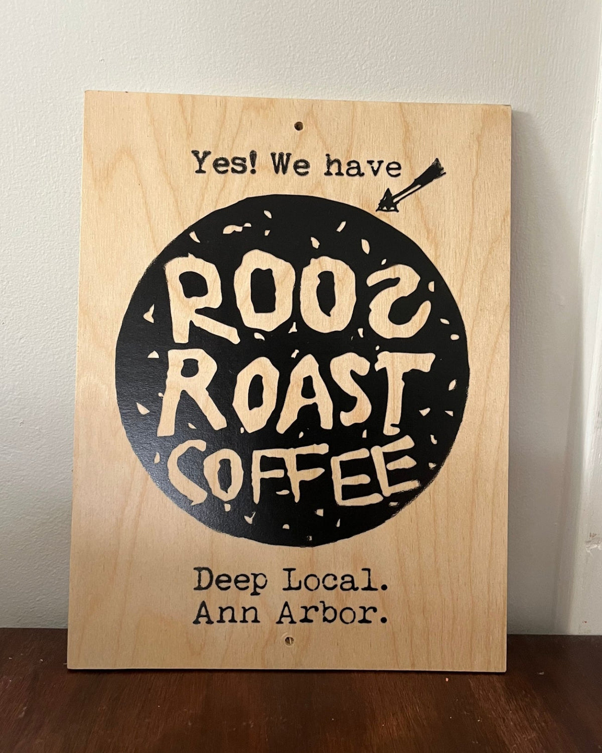 A rectangular wooden sign with a natural light wood finish, featuring a black circular graphic with the words 'ROO2 ROAST COFFEE' in a hand-drawn cream font. The sign also includes the text 'Yes! We have' at the top and 'Deep Local.