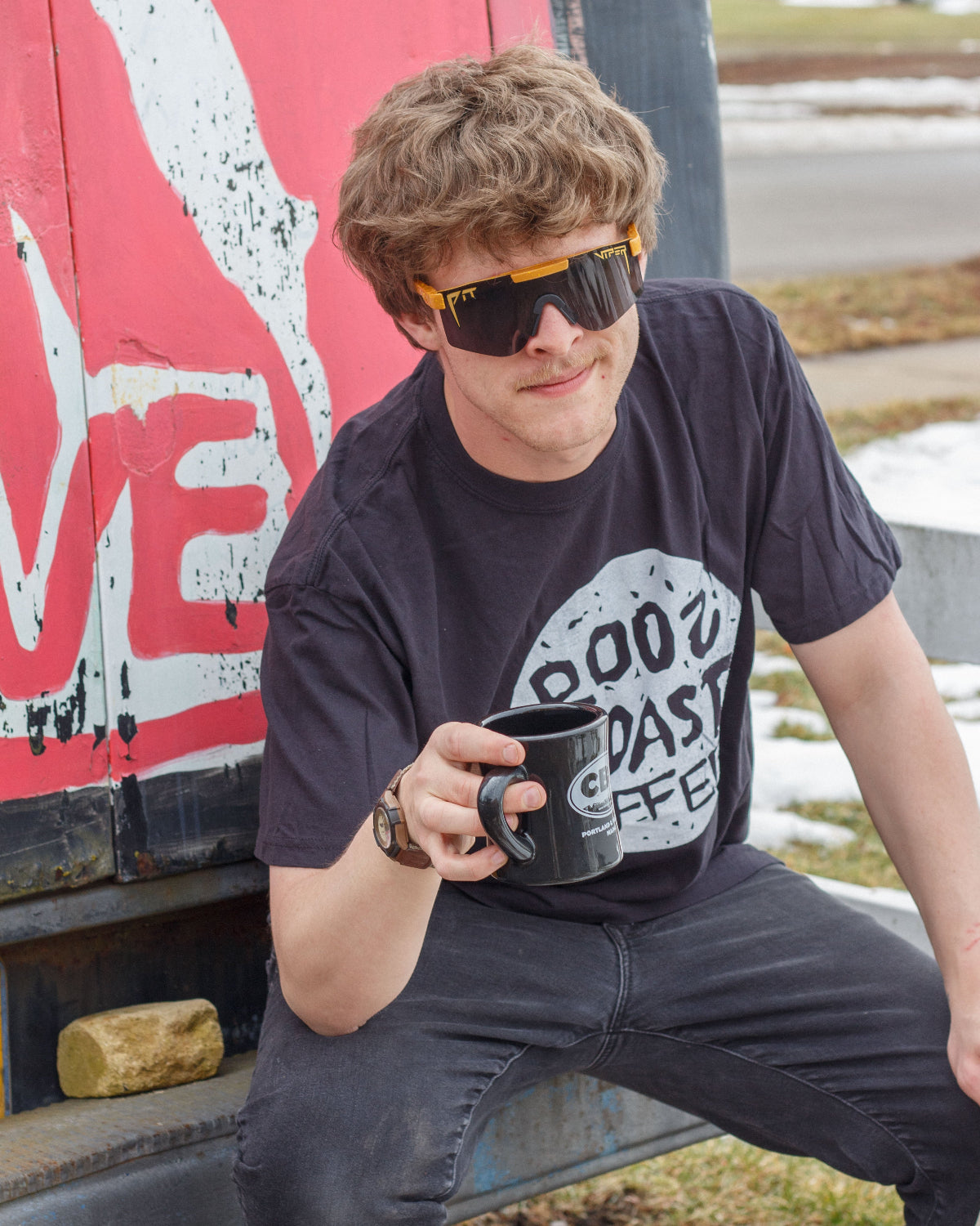 Person holding a coffee mug with a graphic t-shirt, sitting against a graffiti-covered wall.