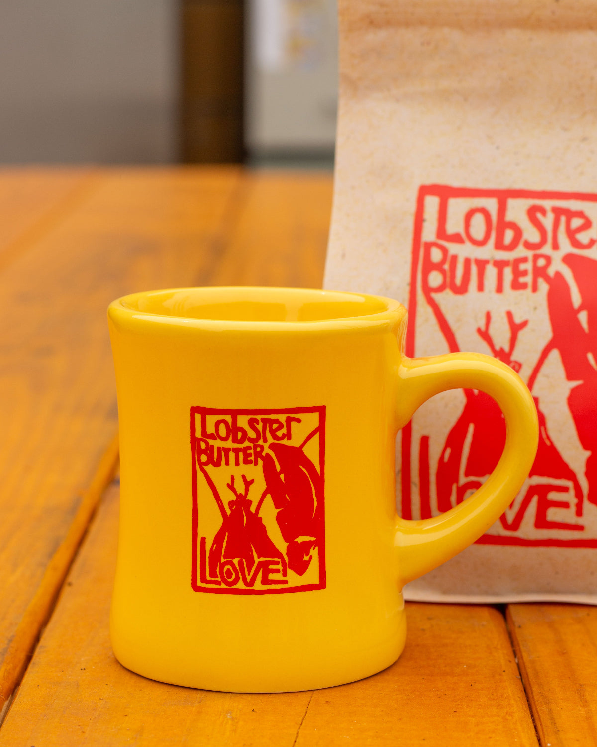 Yellow mug with 'Lobster Butter Love' design on a wooden surface, with a matching bag in the background.