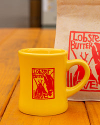 Yellow mug with 'Lobster Butter Love' design on a wooden surface, with a matching bag in the background.
